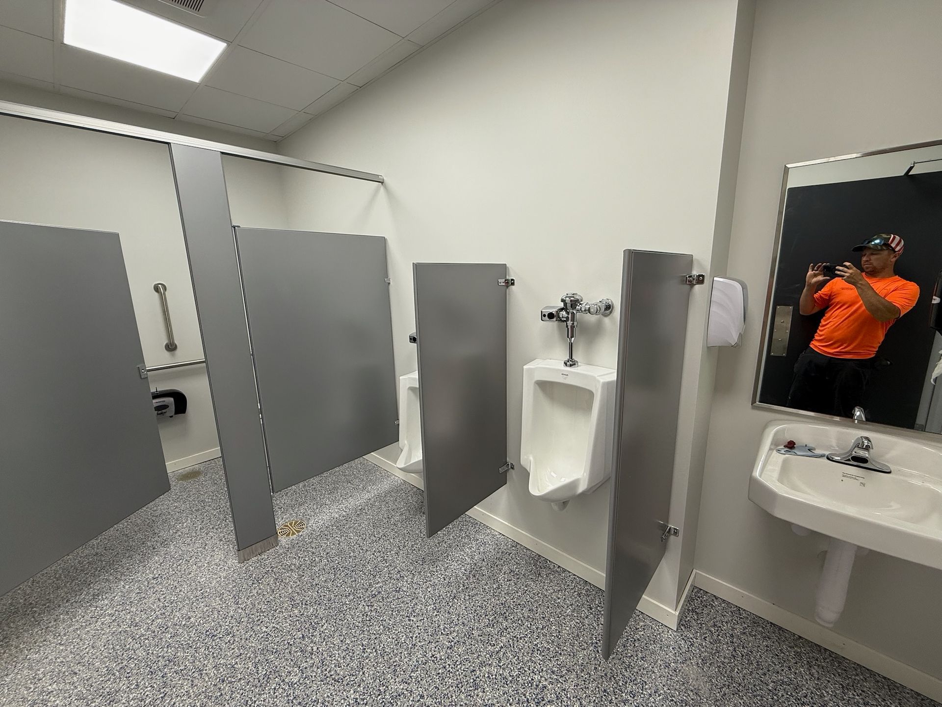 Public restroom with gray stalls, a urinal, and a sink. Man taking a selfie in the mirror.
