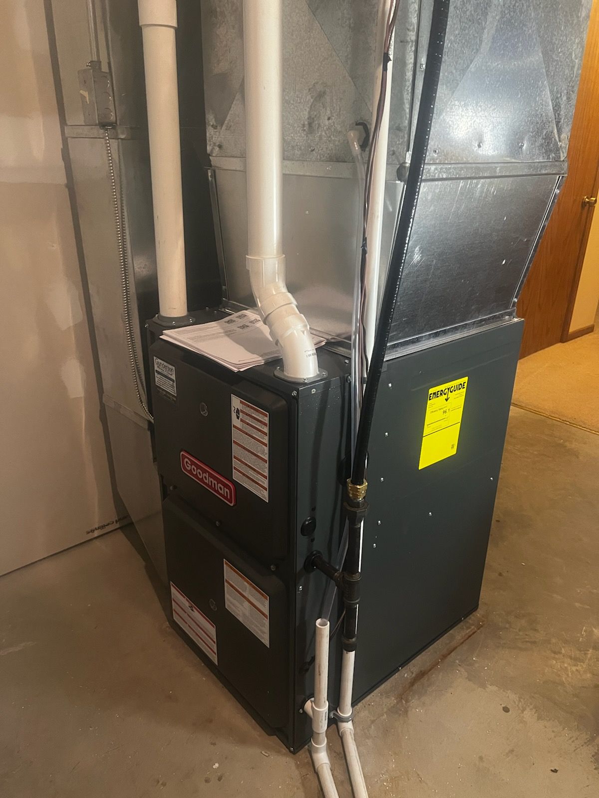 A Goodman furnace in a basement. Gray metal with white PVC pipes.