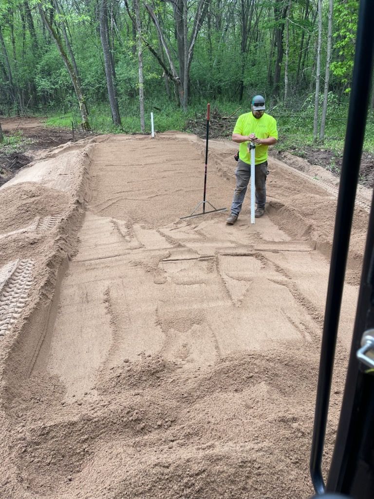 Man with measuring tool in a sand pit, preparing a recreational area in a wooded setting.