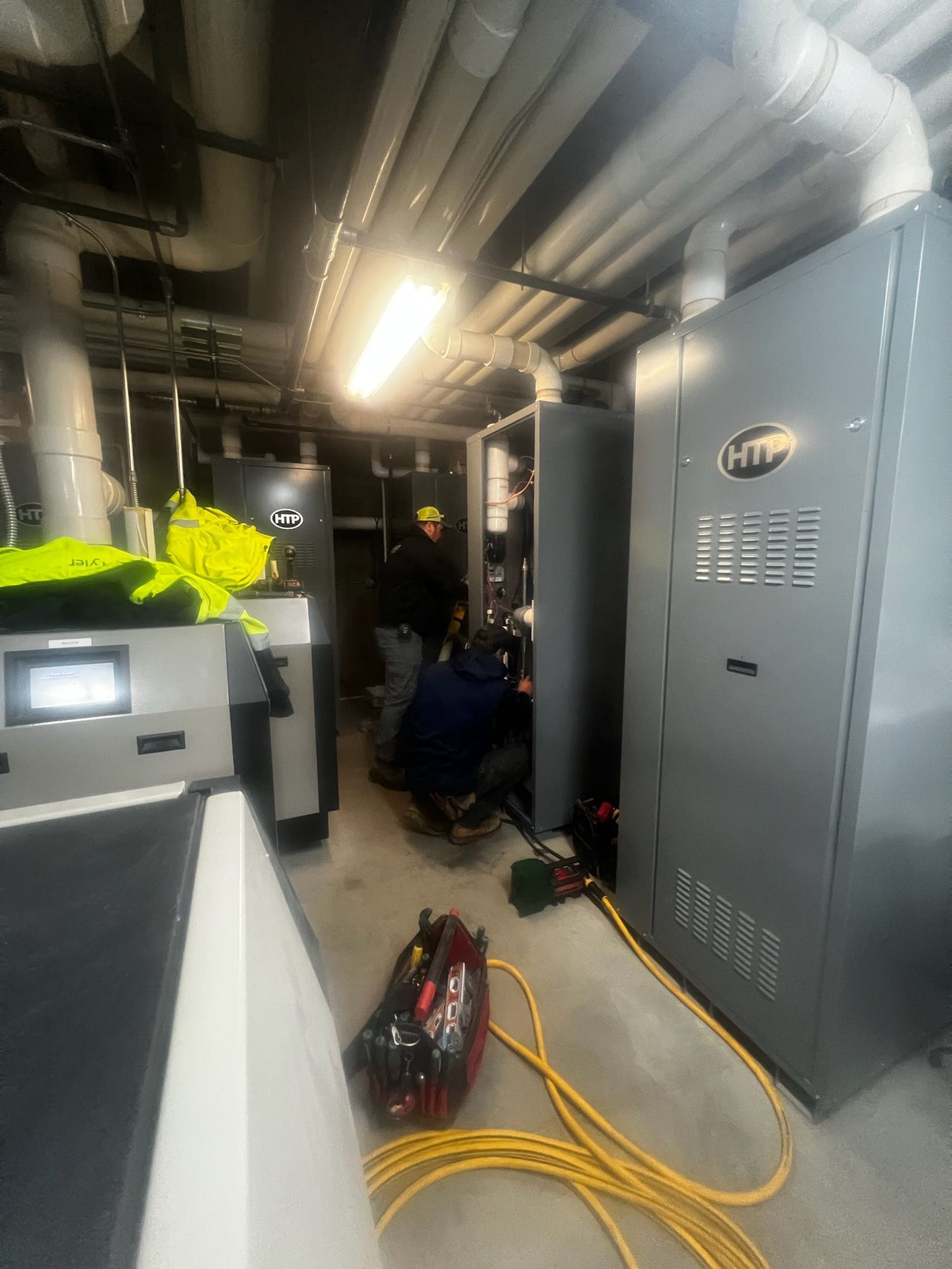 Workers in a utility room installing equipment; large gray machines, tools on the floor, bright lighting, yellow extension cord.