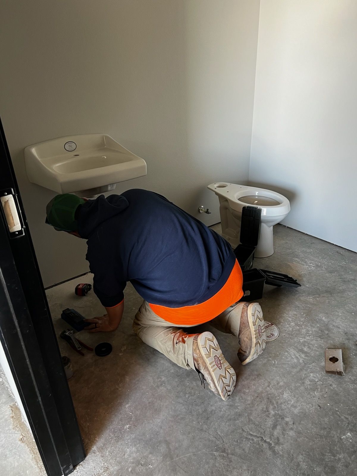 A person crouches in a bathroom working on plumbing; a sink and toilet are present.