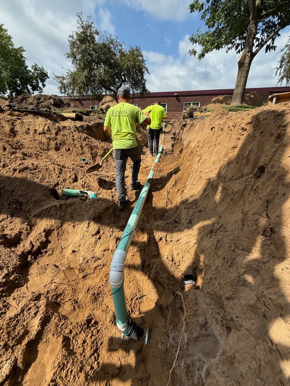 Two workers in neon shirts install a green pipe in a dirt trench under a sunny sky.