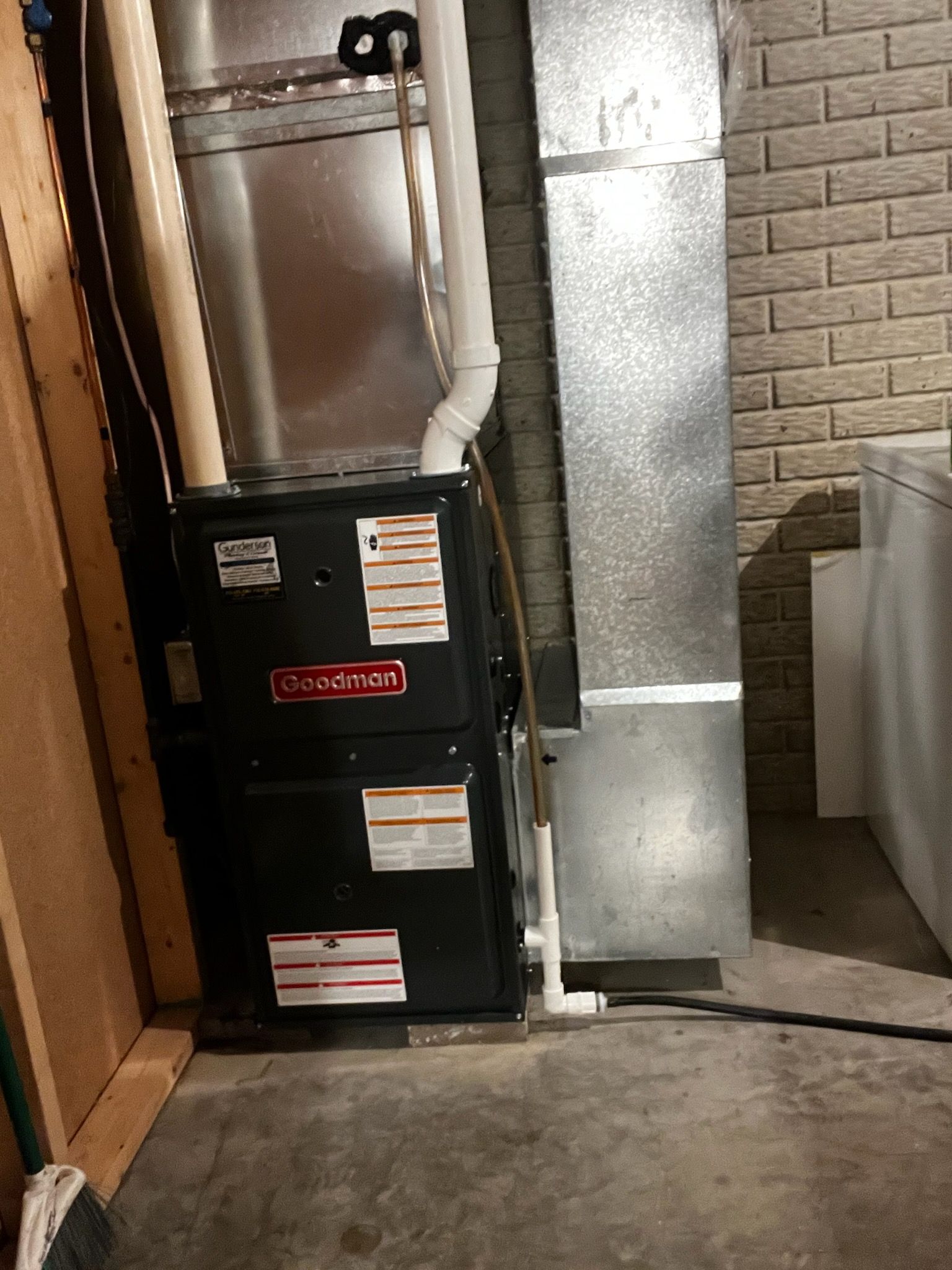 A Goodman furnace in a basement setting.  It's black and silver with white PVC pipes.