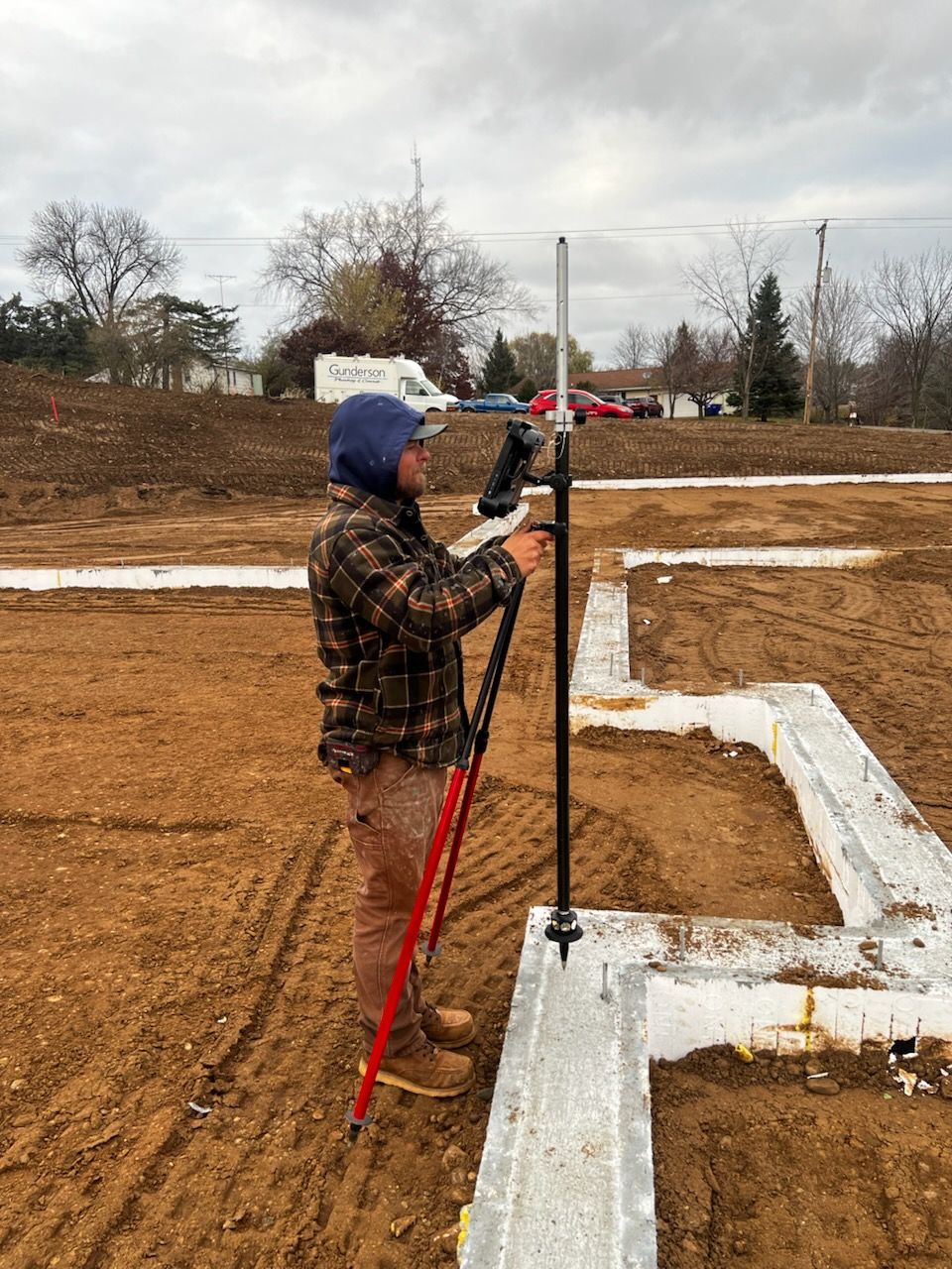 Man using surveying equipment to measure a foundation on a construction site. Brown soil, white foundation, cloudy sky.