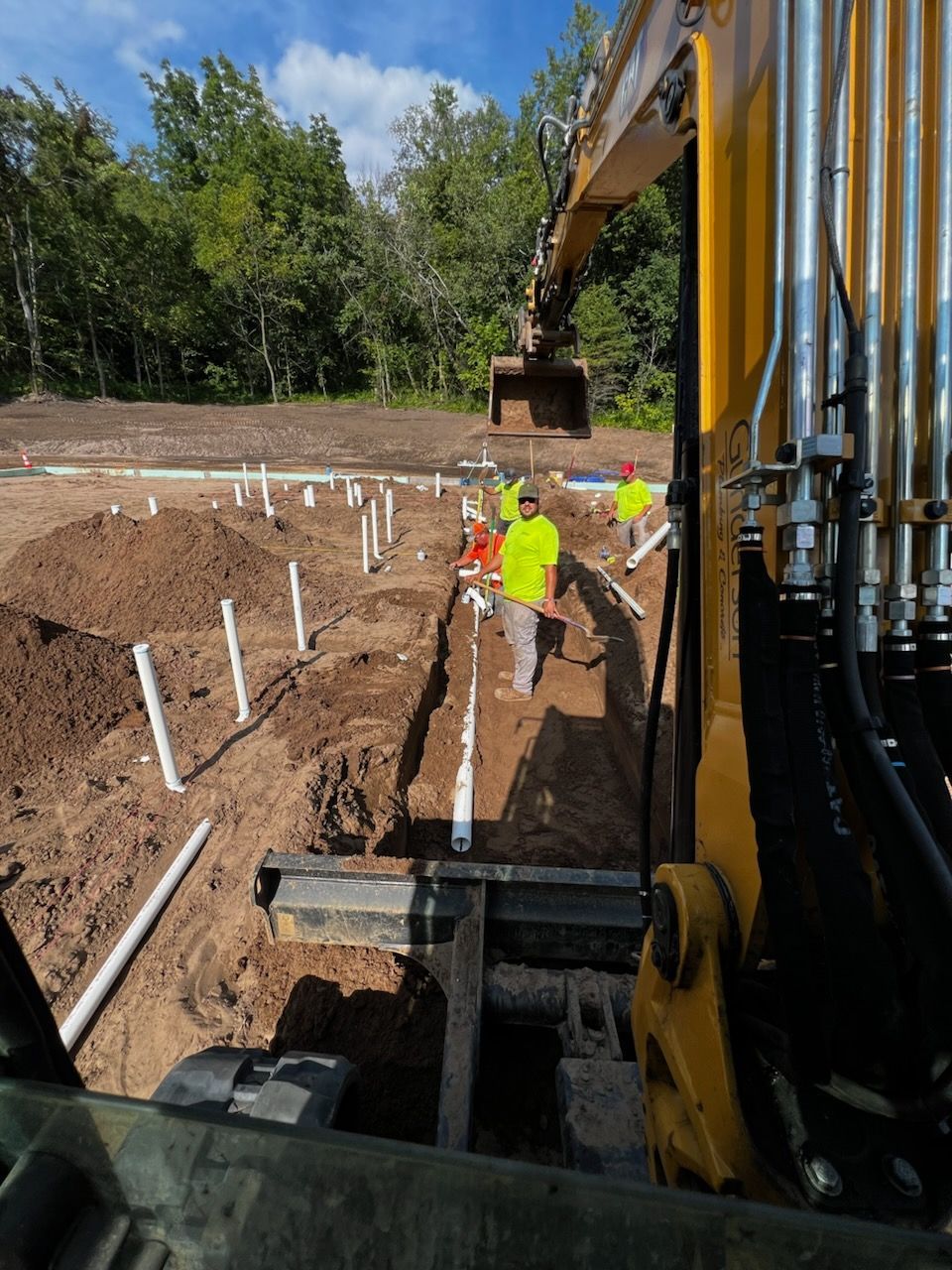 Construction workers installing pipes in a trench, with an excavator in the foreground.