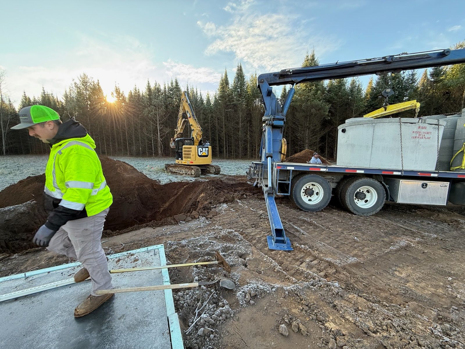 Construction worker in a safety vest walks near a truck unloading concrete blocks with an excavator in the background.