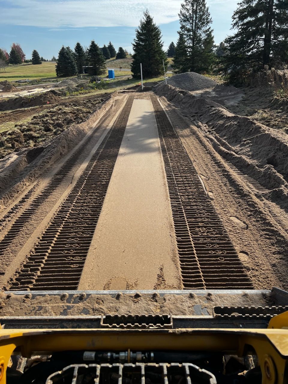 A skid steer machine's view of a level dirt pathway in a construction site, under a blue sky.