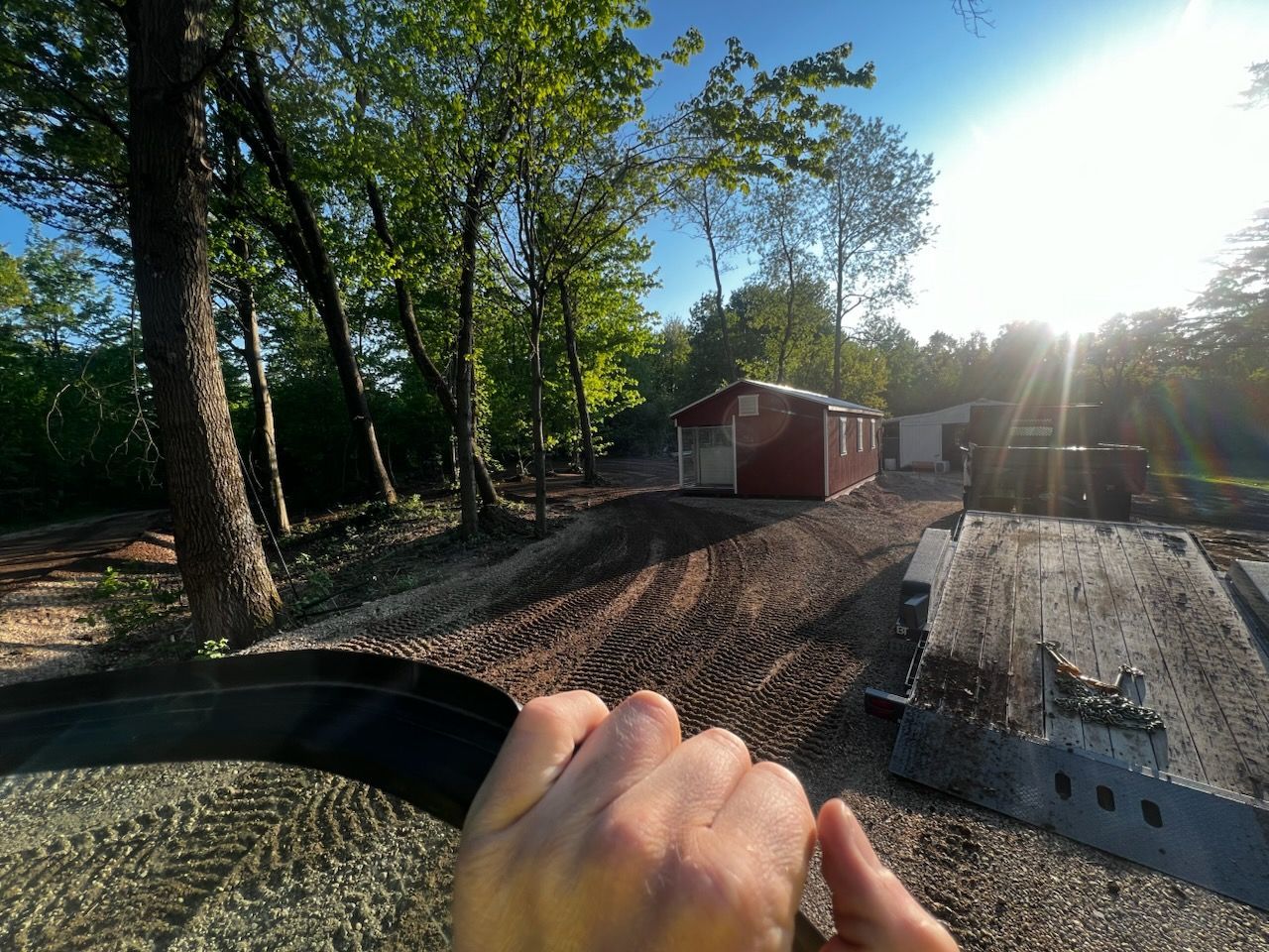 Hand on machinery controls, view of a red building in a wooded area with bright sunlight.