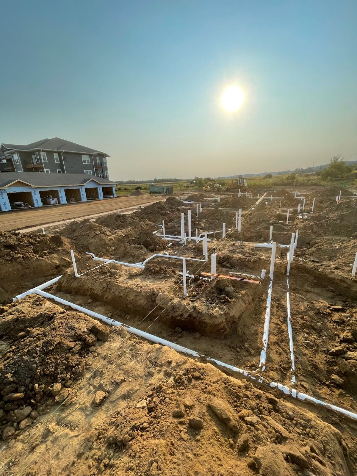 Construction site with white pipes in trenches; apartment building in the background under a sunny sky.