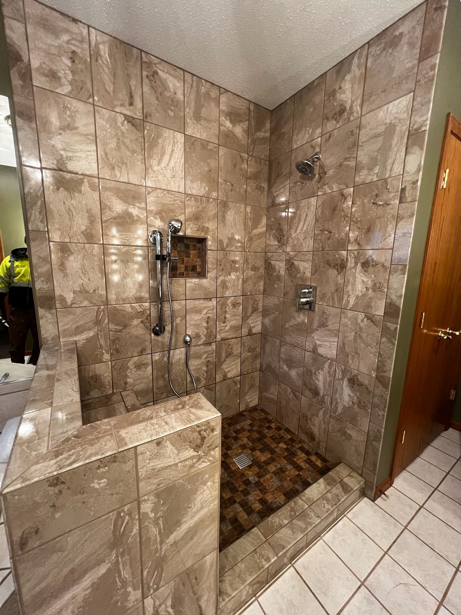 Walk-in shower with tan tile walls and floor, multiple shower heads, and a small brown tiled barrier.
