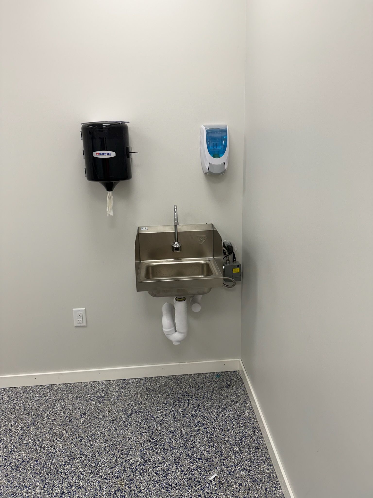 Stainless steel sink with paper towel and soap dispensers on a gray wall. Floor is speckled.
