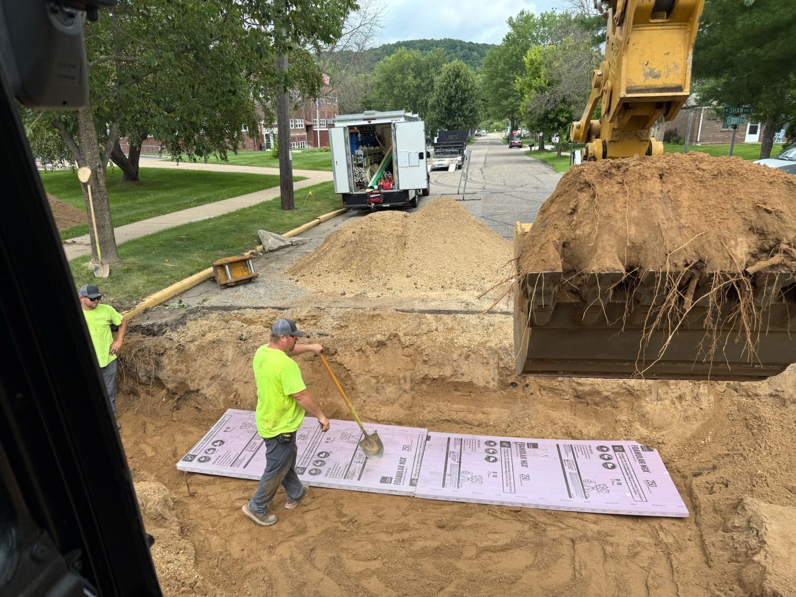 Workers install pink insulation sheets in an excavation pit.  An excavator and utility truck are visible.