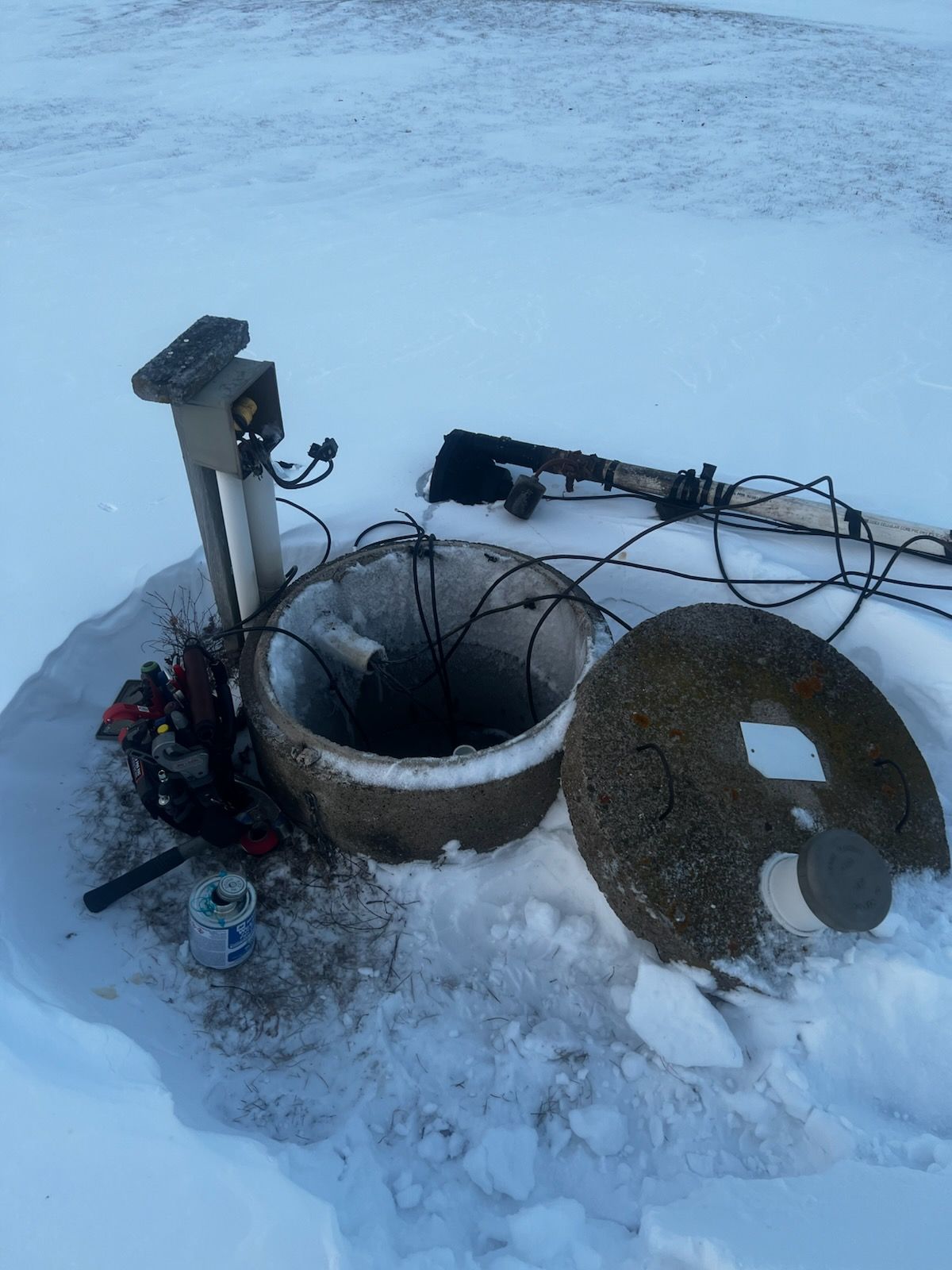 Open concrete well with lid removed in snow, electrical components visible.