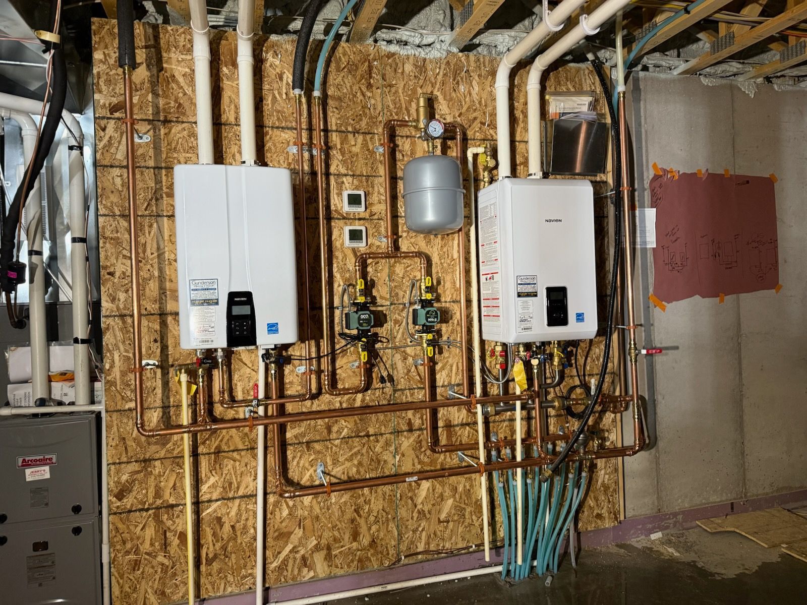 Two white tankless water heaters with copper pipes and plumbing in a basement.