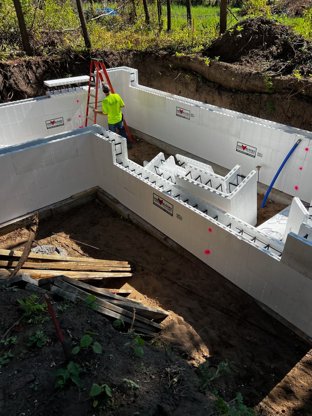 Construction site with white ICF walls and a worker in neon shirt standing near red ladder.