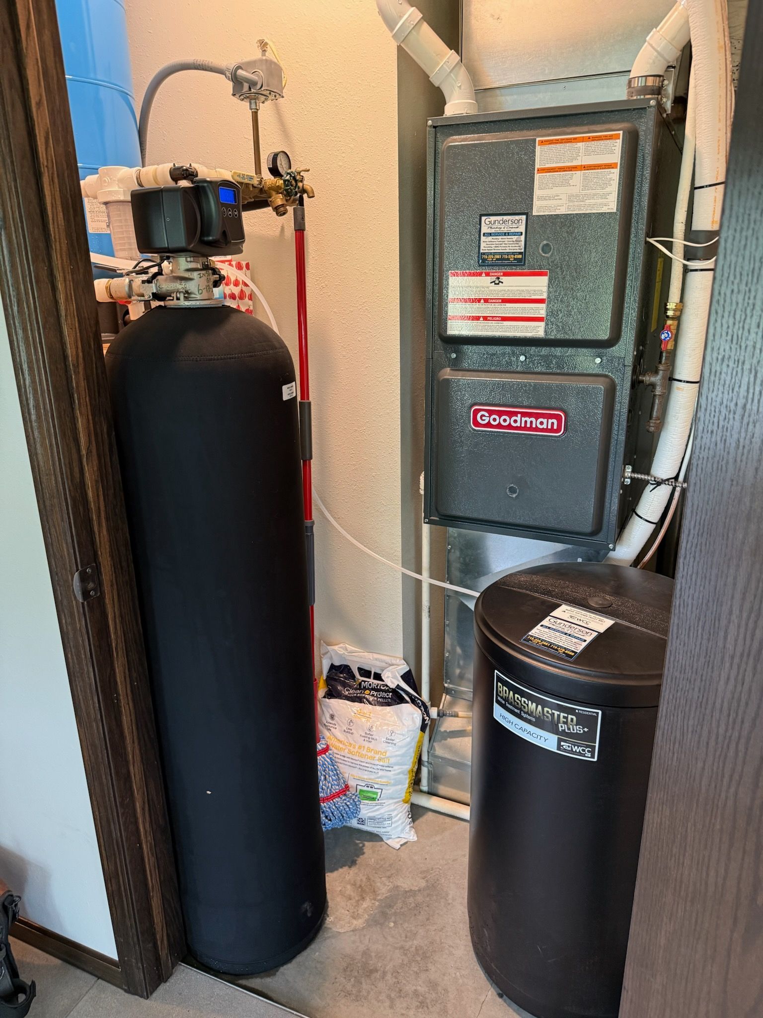 Water softener system and furnace in a utility room. Black tanks, metal furnace, white piping.