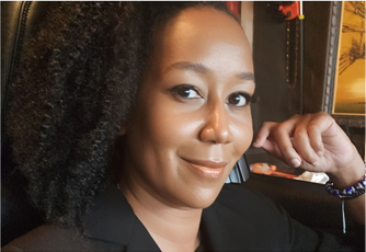 Woman smiling, leaning in, arm at cheek, wearing black jacket, dark curly hair.