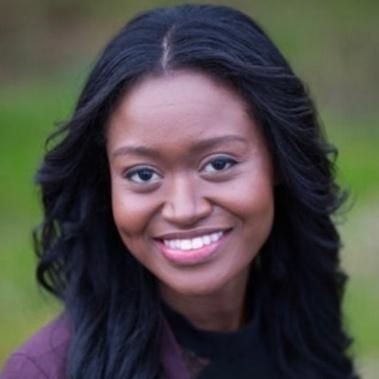 Woman with long, dark hair smiles outdoors, wearing a dark top and burgundy jacket; green background.