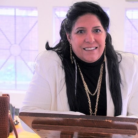 Woman in white blazer and black turtleneck smiles at a lectern with a window behind her.