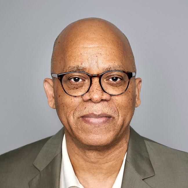 Bald man wearing glasses, khaki jacket, smiling in front of a neutral gray background.