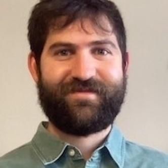 Man with dark hair and beard, wearing a blue button-down shirt, smiling.