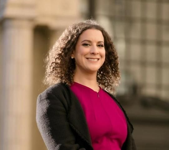 Woman with curly hair smiles, wearing a pink top and black jacket, in front of a building with columns.