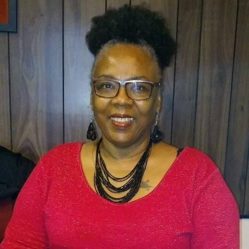 Woman wearing glasses, a red shirt, and black jewelry smiles in a wood-paneled room.