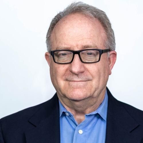 Man with glasses in blue shirt and navy blazer smiles against white backdrop.