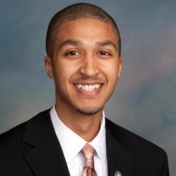 A man in a suit smiles, wearing a tie; studio portrait.