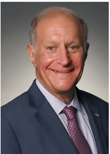 Smiling man in a blue suit and purple tie, against a gray background.