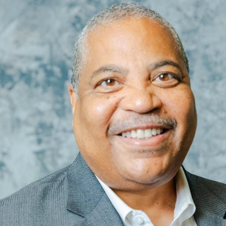 Man with short grey hair and mustache smiling, wearing a gray blazer and white shirt.