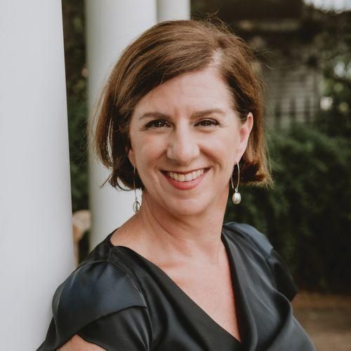Woman with short brown hair smiles, leans against a white column, wears a dark top, and earrings.