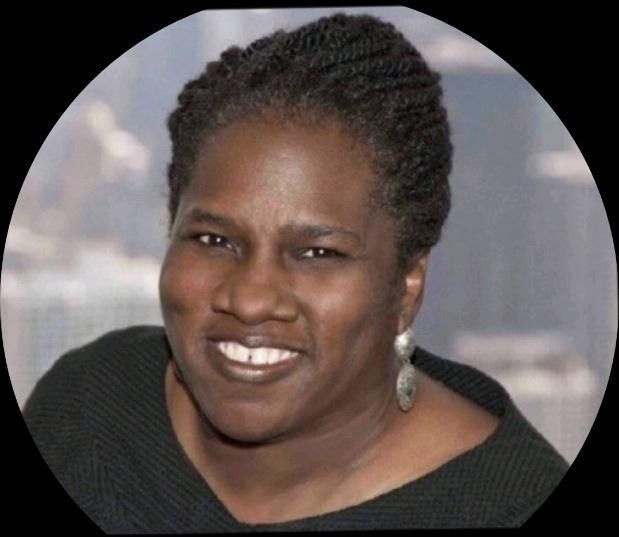 Woman with short, dark braided hair, smiling, wearing large earrings and a black top. Blurred cityscape in background.