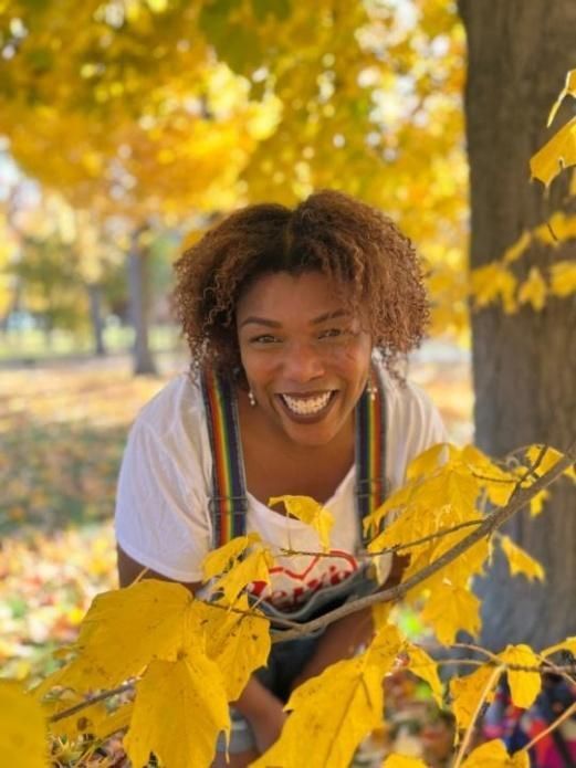 Woman smiling, framed by yellow autumn leaves; wears white shirt and overalls in park setting.