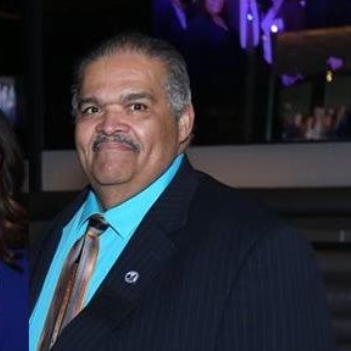 Man in blue shirt, striped tie, and dark suit smiling, in a brightly lit setting.