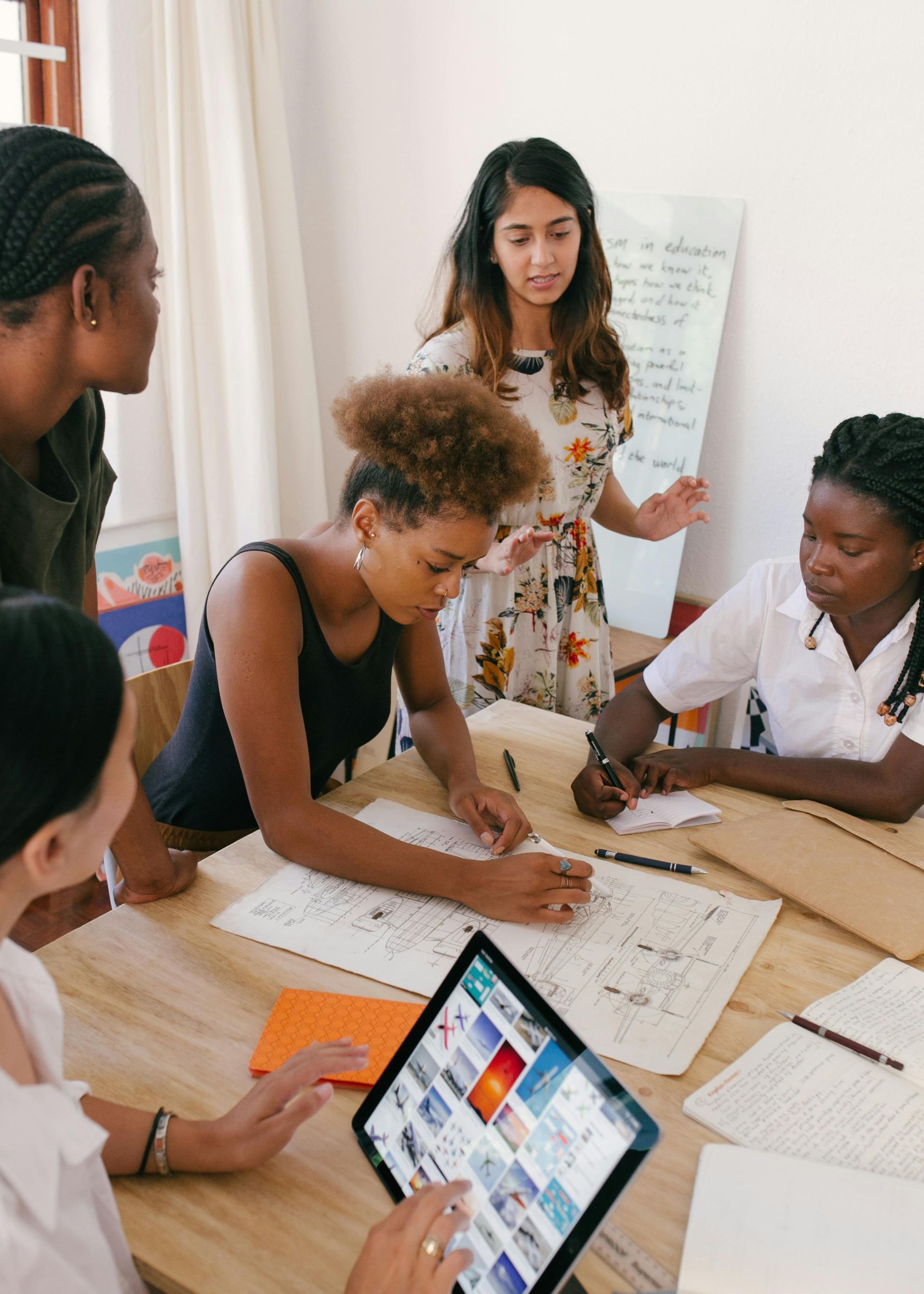 A group of people collaborating at a table, using a tablet and papers. One person is gesturing to a whiteboard.