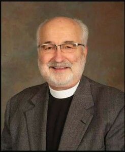 Portrait of a smiling clergyman in a gray suit and black clerical shirt with white collar.