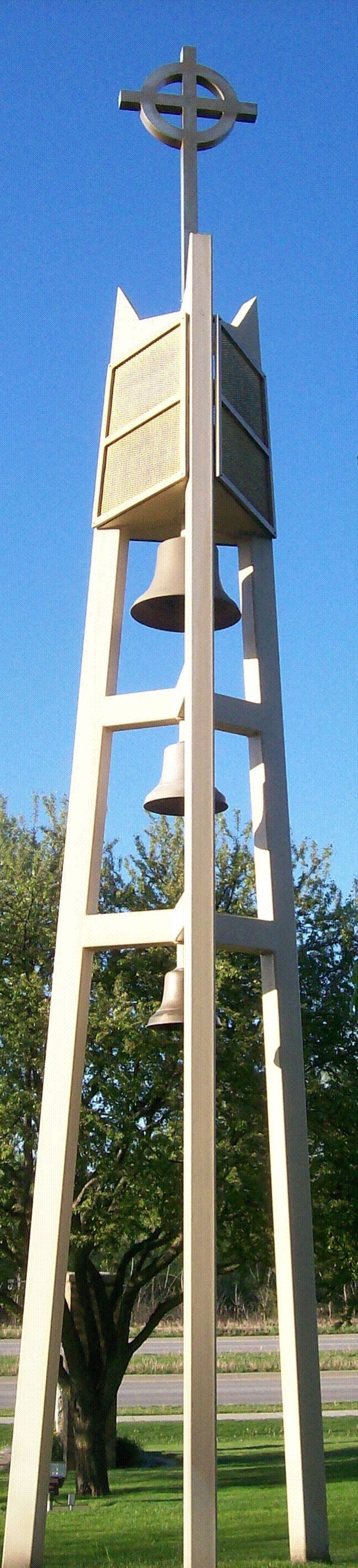 Tall wooden tower with a cross-like structure at the top against a blue sky and trees