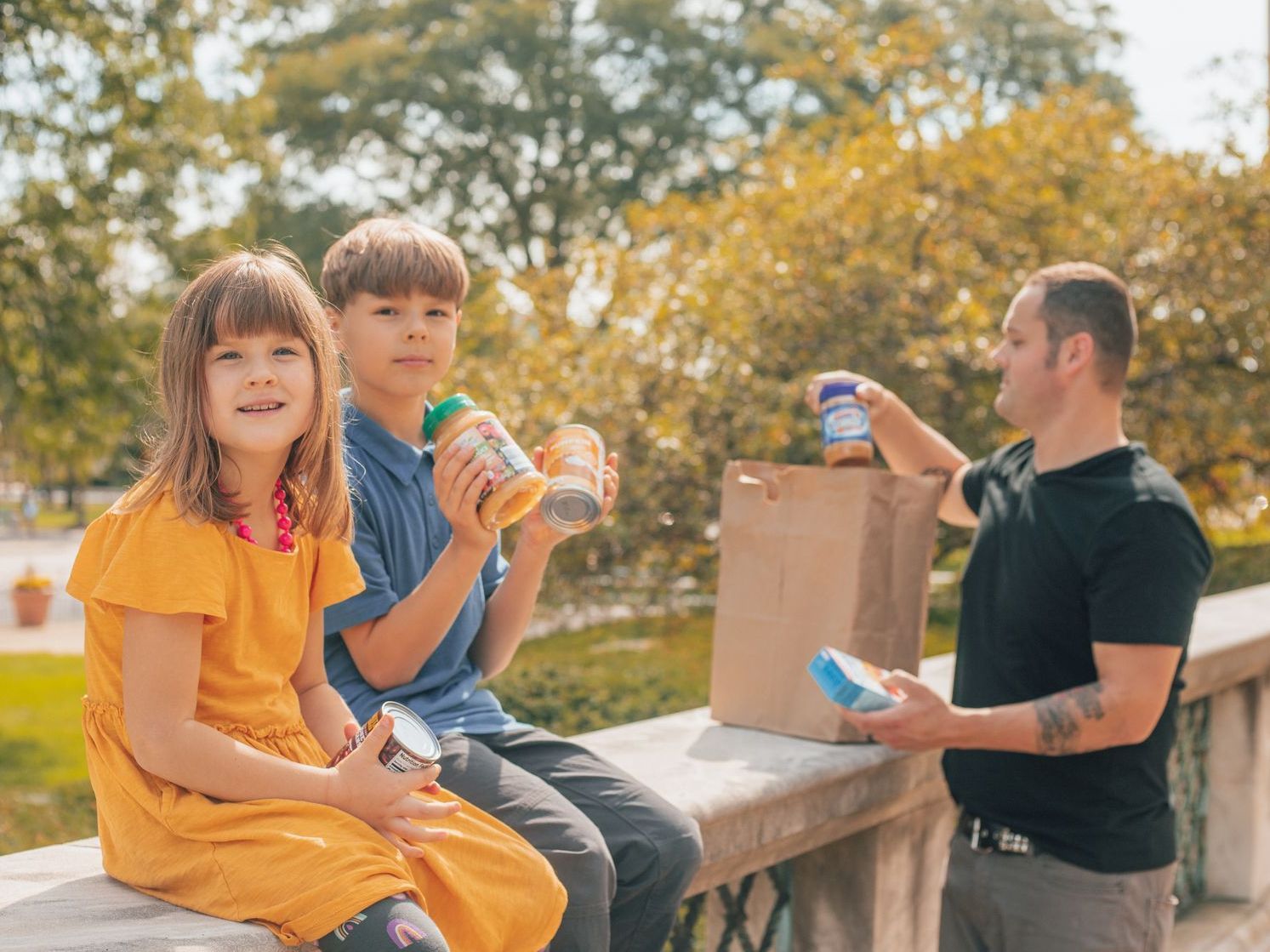 Two children and a man share snacks outdoors beside a paper bag on a sunny day.