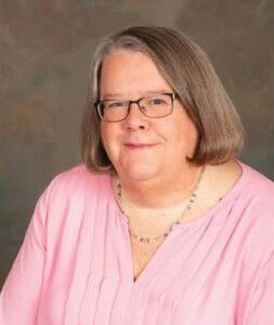 Portrait of a person with glasses and shoulder-length hair, wearing a pink blouse and necklace, smiling gently