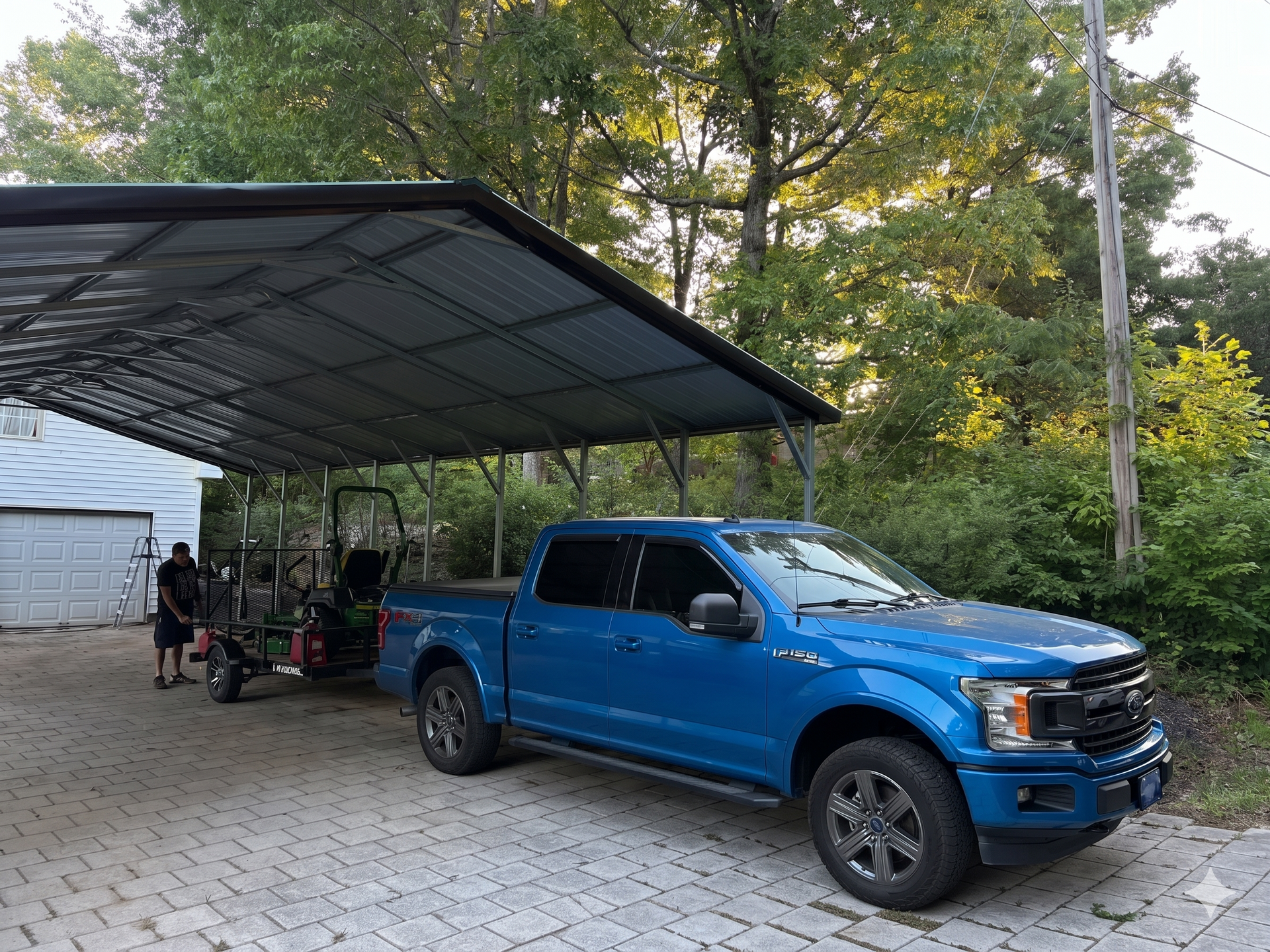 A blue Ford truck parked under a metal carport, towing a lawnmower on a trailer, with a man standing nearby.