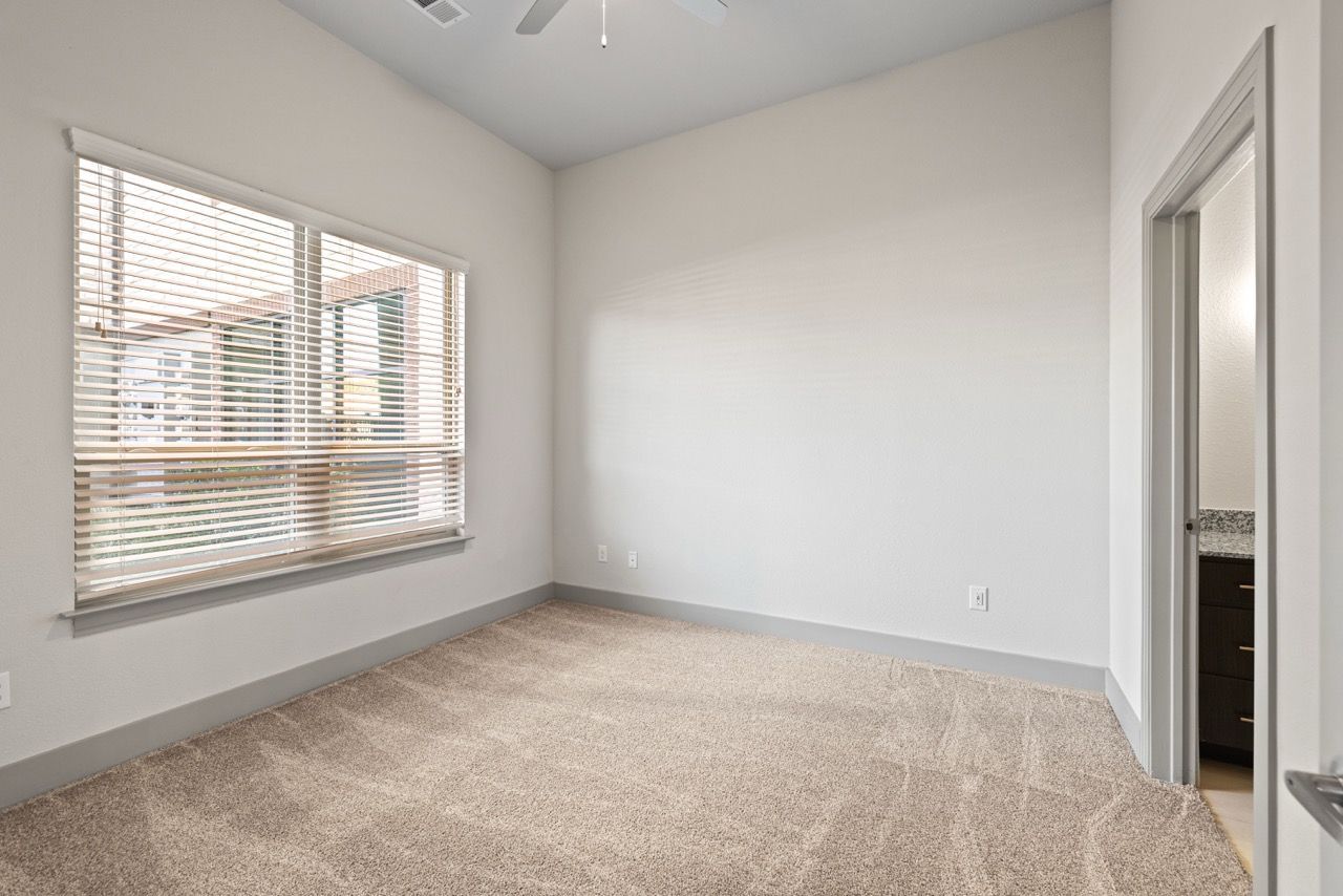 Interior of an unfurnished apartment room with a large window, blinds, and beige carpet.