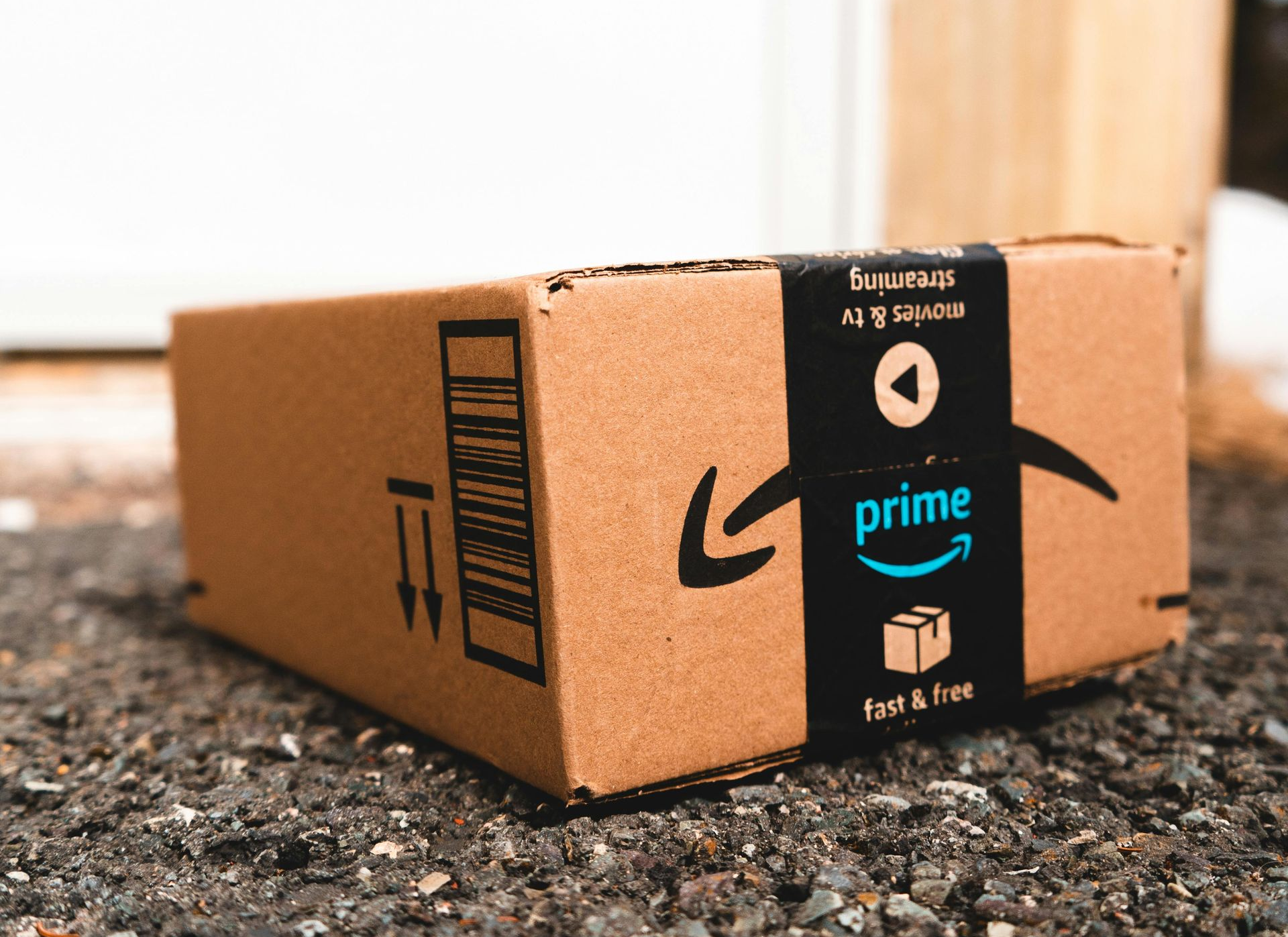 An Amazon Prime cardboard package sits on a gravel driveway in front of a house door.