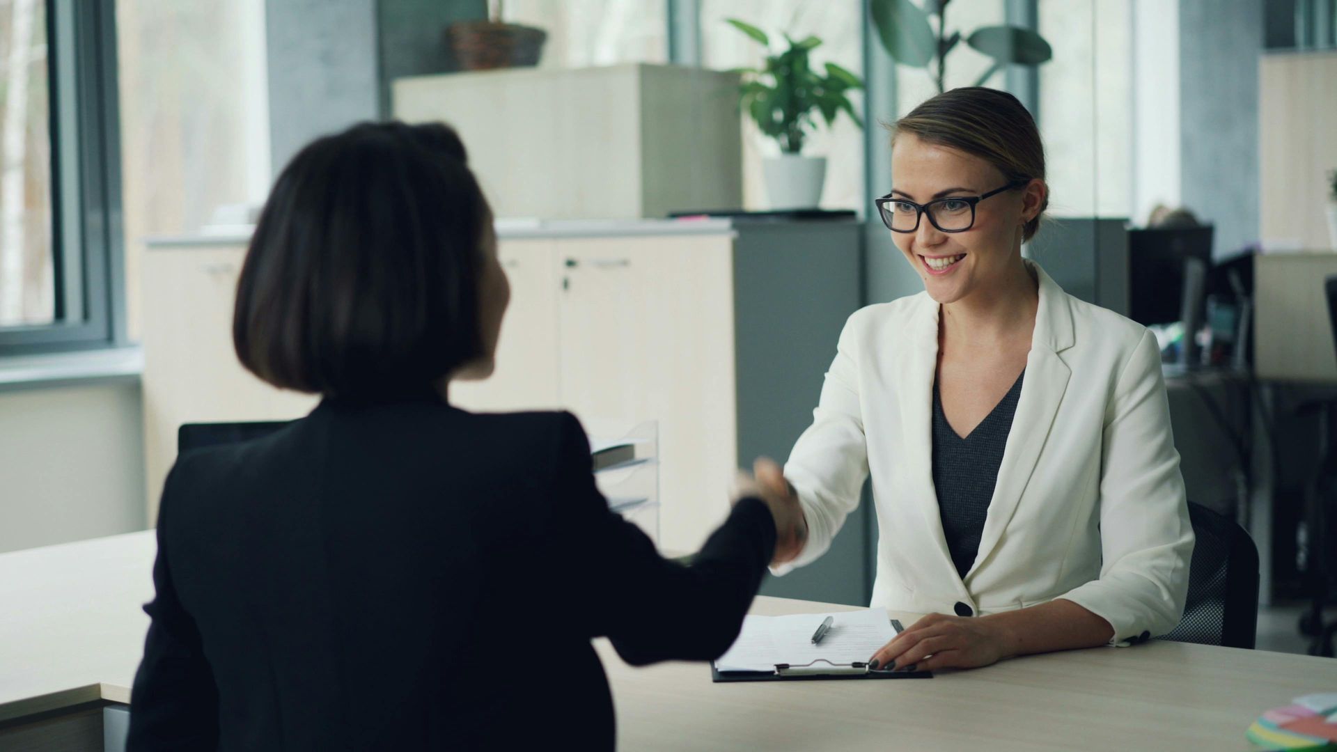 Two professionals shake hands over a desk in a bright office, one smiling while the other is seen from behind.