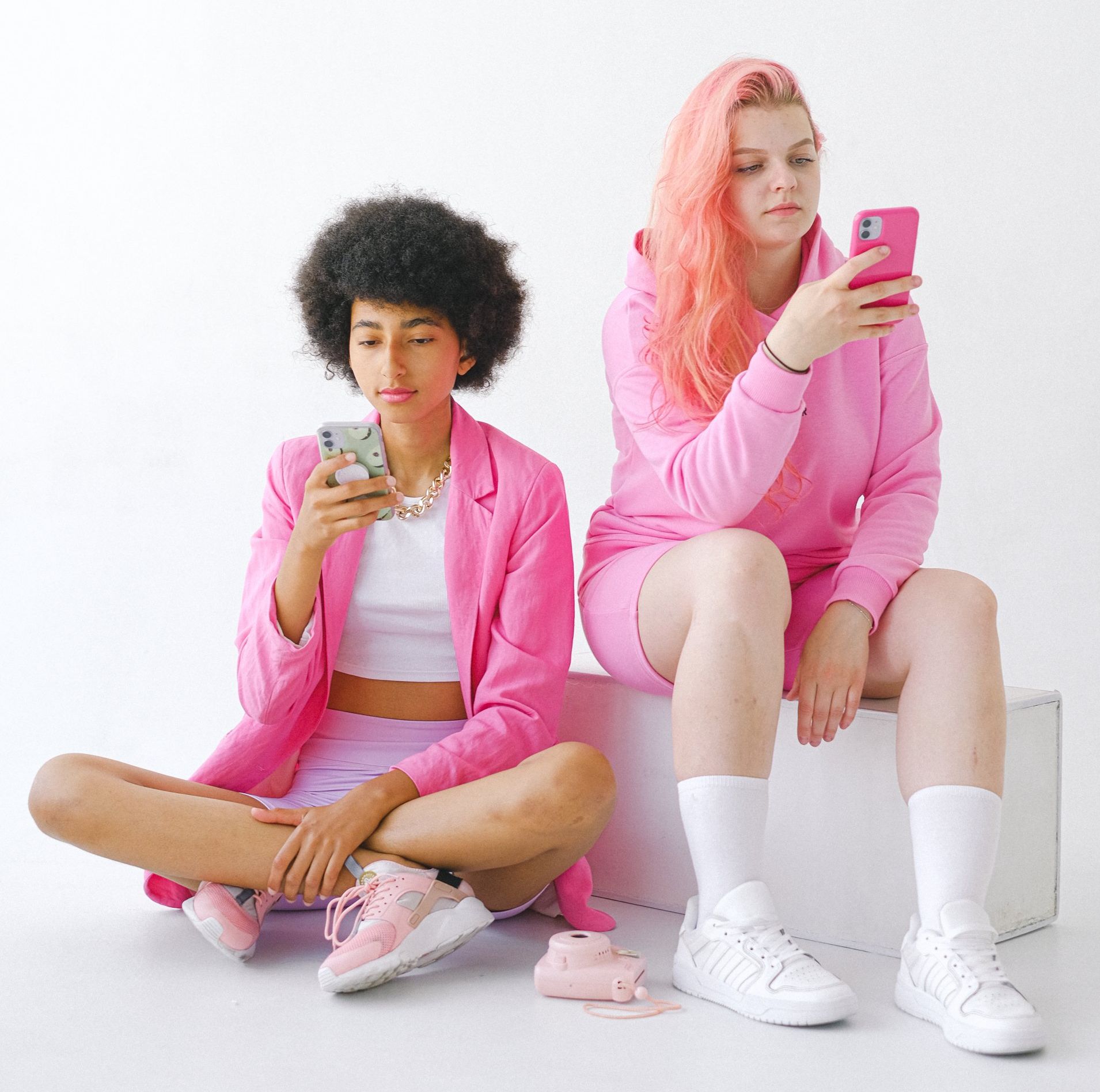 Two women in pink clothes are sitting on the floor looking at their phones.