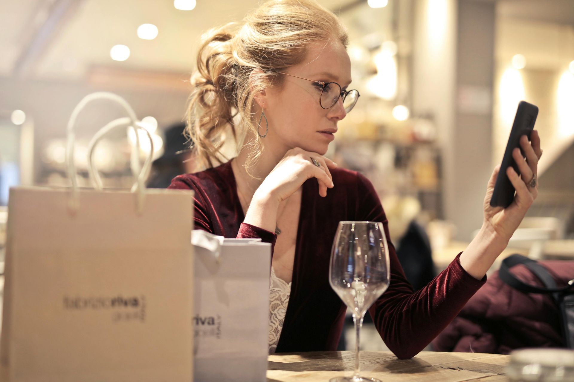 A woman is sitting at a table with a glass of wine and looking at her phone.