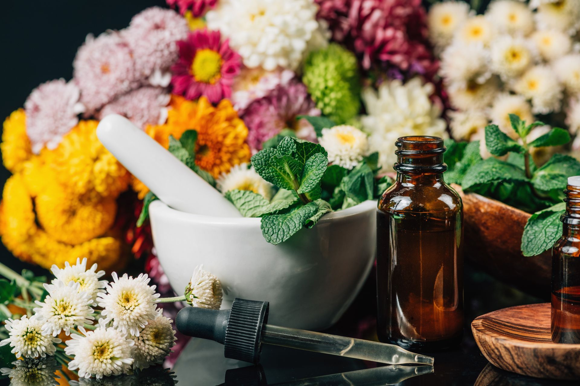 Flowers, mortar, pestle, essential oil bottles, and herbs, arranged on a dark surface.