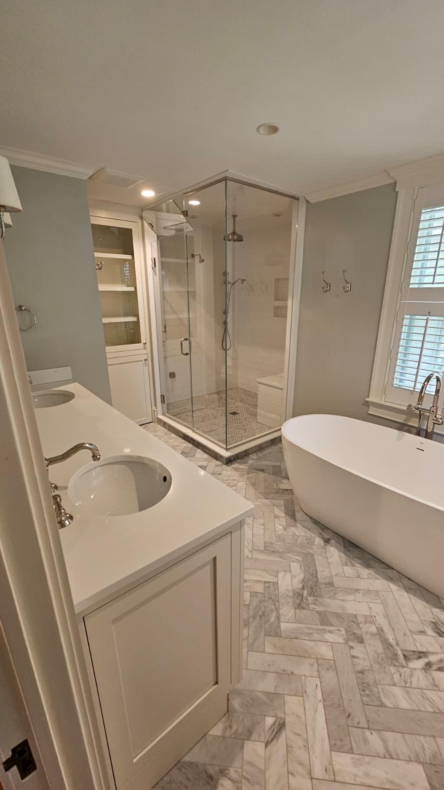 A bright bathroom with a white freestanding tub, a glass shower, a vanity with a sink, and herringbone marble floors.