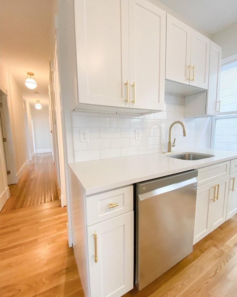 A bright kitchen with white cabinets, gold hardware, a stainless steel dishwasher, and a view into a hallway.