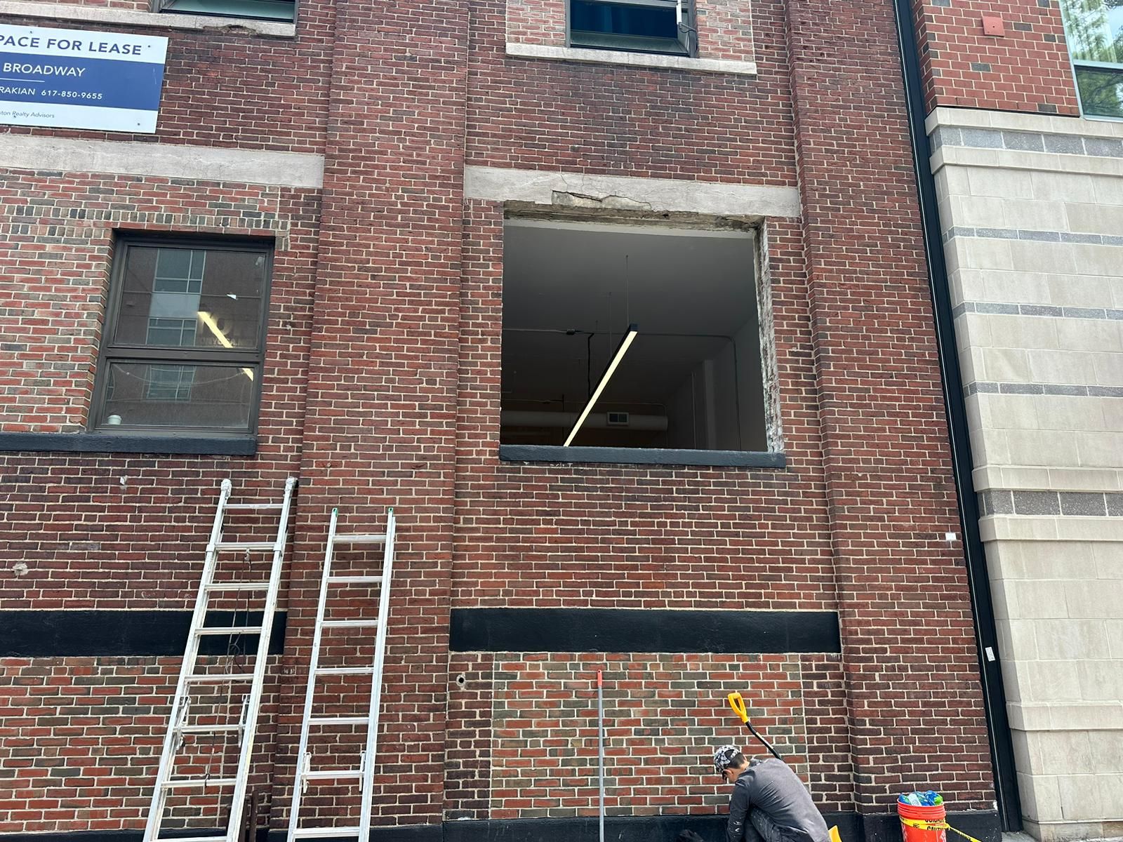 Brick building exterior under renovation with two metal ladders leaning against the wall and a worker below.